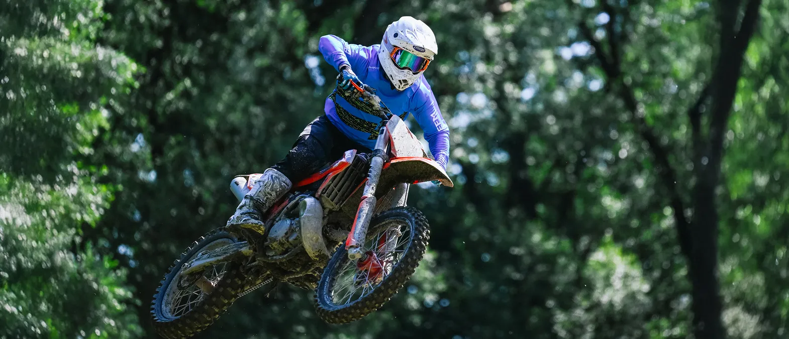 Ein Motorradfahrer in blauer, langer Rennbekleidung und weißem Helm springt mit seinem Motorrad durch einen Wald.