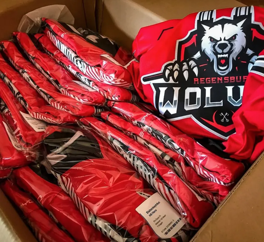 Eine Kartonbox mit mehreren roten, individuell bedruckten Sporttrikots, die in Plastikfolie verpackt sind, eines davon mit einem Wolf-Logo und der Aufschrift 'REGENSBURG WOLVES'.