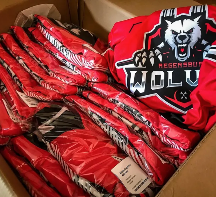 Eine Kartonbox mit mehreren roten, individuell bedruckten Sporttrikots, die in Plastikfolie verpackt sind, eines davon mit einem Wolf-Logo und der Aufschrift 'REGENSBURG WOLVES'.