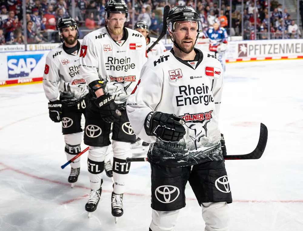 Drei Eishockeyspieler in weißen Trikots mit schwarzem Logo und grafischen Designs stehen auf dem Eis.