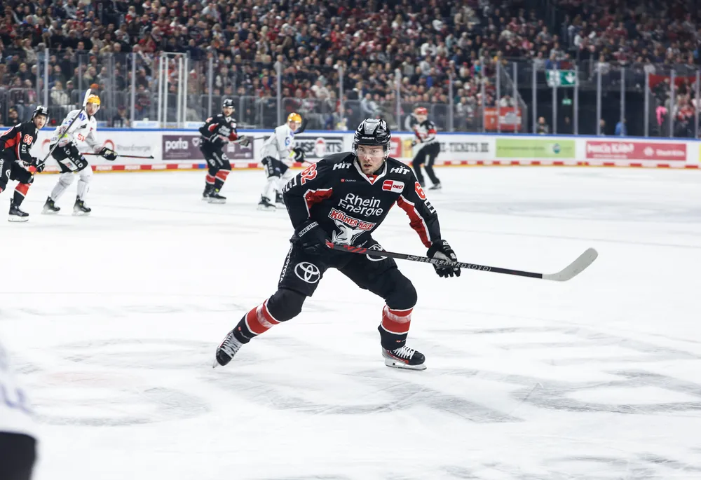 Ein Eishockeyspieler in einem schwarzen Trikot mit roten und weißen Akzenten, der mit einem Schläger auf dem Eis spielt.