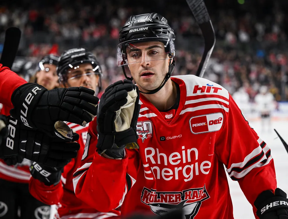 Ein Eishockeyspieler trägt ein rotes Trikot mit weißen und schwarzen Details, während er mit einem Teamkollegen interagiert.