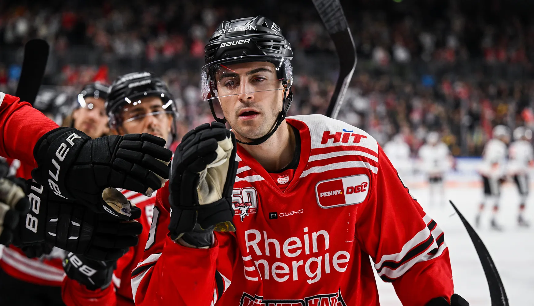 Ein Eishockeyspieler in einem roten Trikot mit weißen Streifen und Handschuhen, der auf das Spiel reagiert.