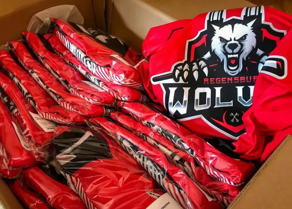 Ein Karton mit roten, individuell bedruckten Sporttrikots, darunter ein Trikot mit einem Wolf-Logo und der Aufschrift 'Regensburg Wolves'.