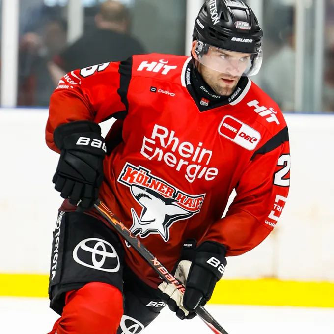 Ein Eishockeyspieler in einer roten Trikot mit dem Logo von Rhein Energie und Kölner Haie, der auf dem Eis steht.
