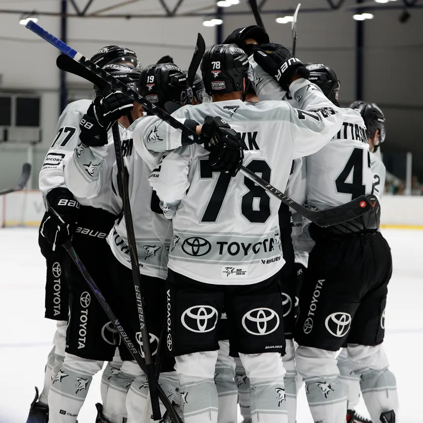 Eine Gruppe von Eishockeyspielern in weißen Trikots umarmt sich auf dem Eis nach einem Tor.