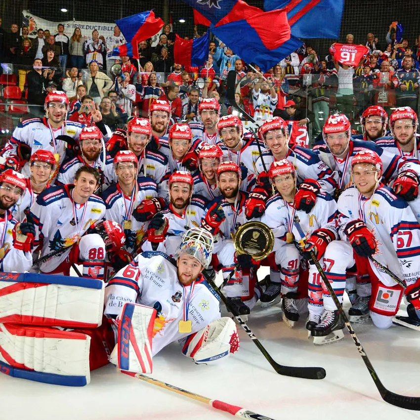 Eine Gruppe von Eishockeyspielern in weißen Trikots mit roten und blauen Akzenten posiert stolz mit Medaillen und einer Trophäe auf dem Eis.