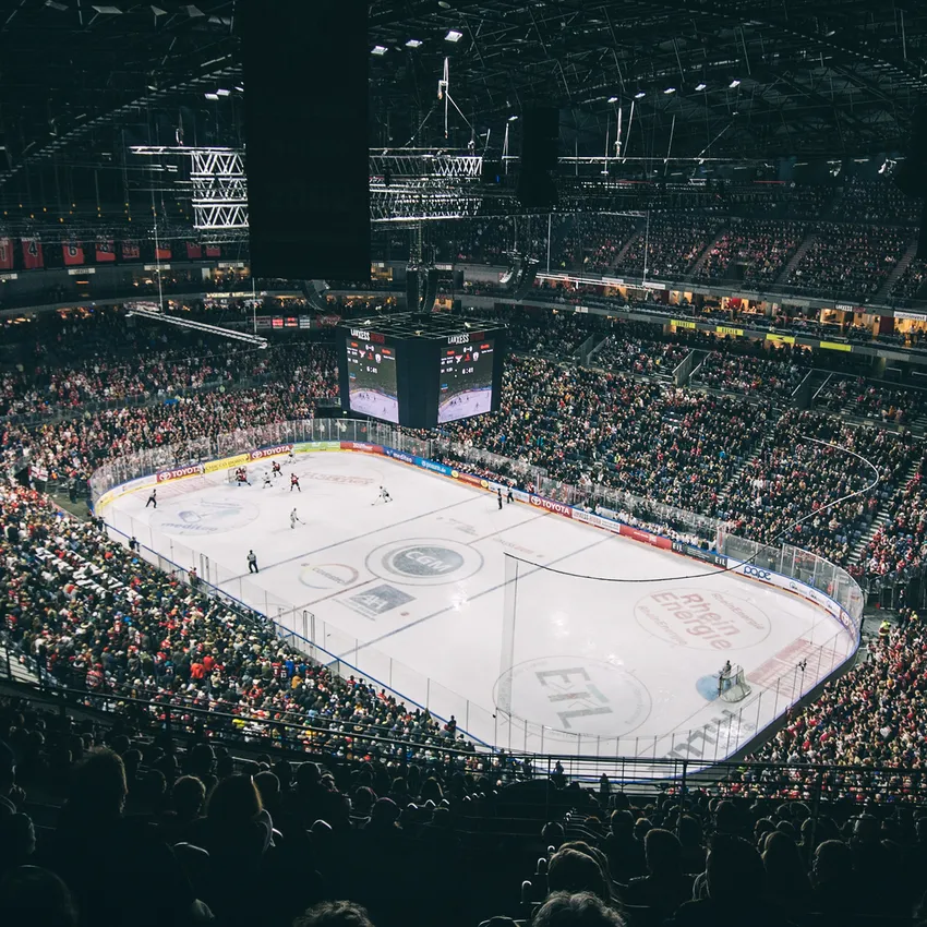 Eine vollbesetzte Eishockey-Arena während eines Spiels, mit Spielern auf dem Eis und einer großen Anzeigetafel im Hintergrund.