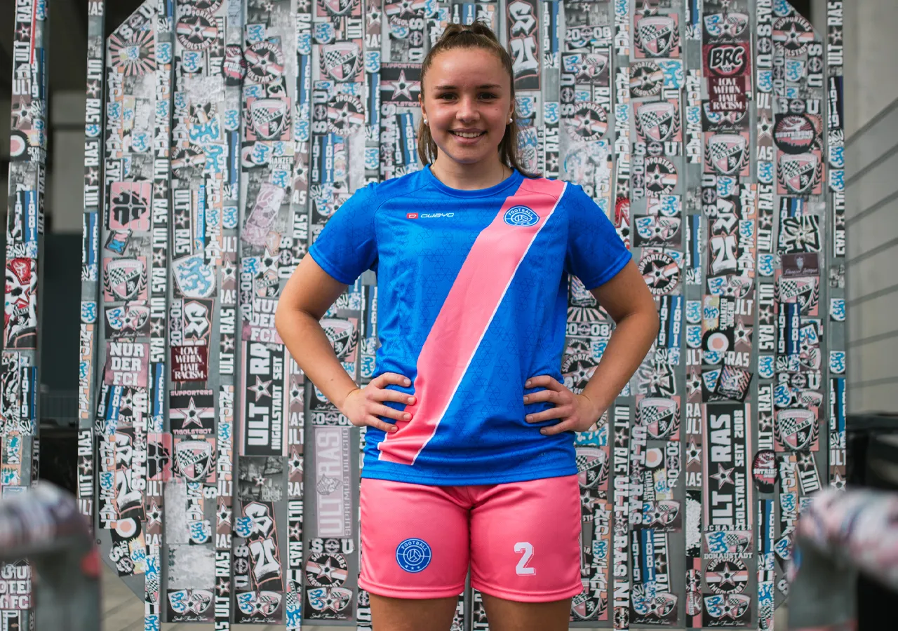 Eine junge Frau trägt ein blaues Fußballtrikot mit einer pinken diagonalen Linie und pinken Shorts mit der Nummer 2, vor einer Wand mit Grafiken und Aufklebern.