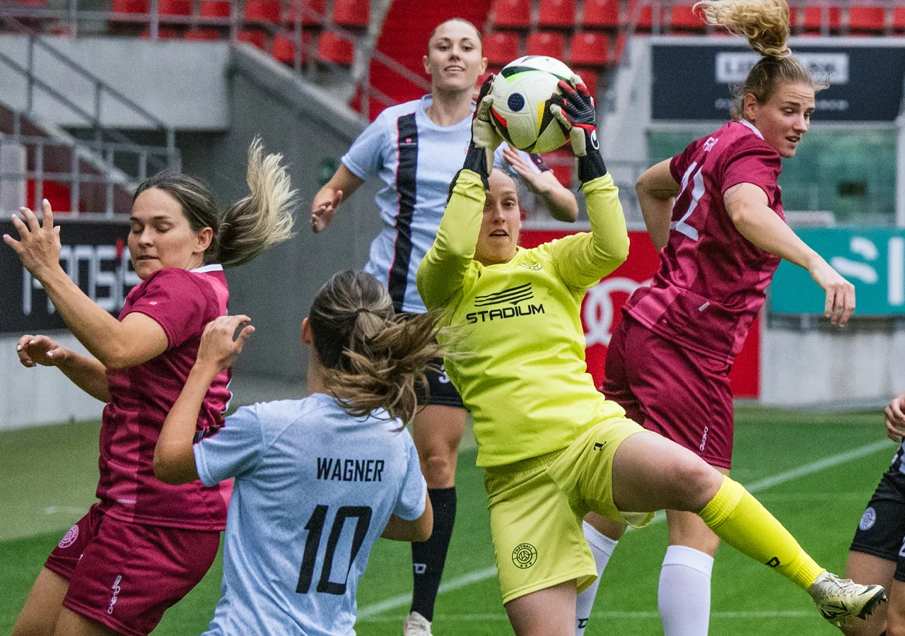 Eine Torhüterin in gelber Sportbekleidung fängt einen Fußball, während andere Spielerinnen in verschiedenen Trikots um den Ball kämpfen.