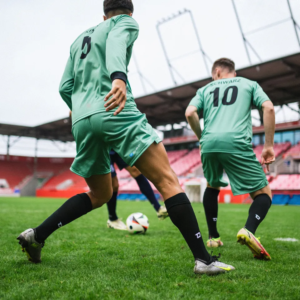 Zwei Fußballspieler in grünen Trikots und schwarzen Socken, die auf einem Rasenfeld in einem Stadion aktiv spielen.