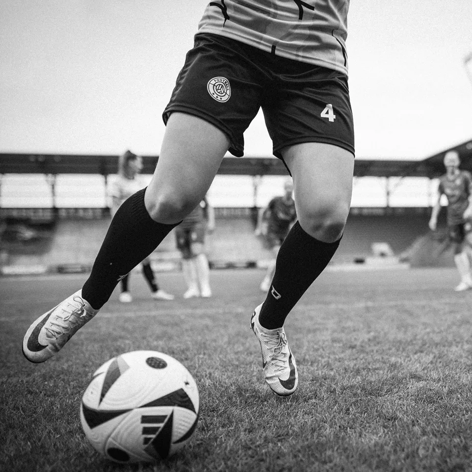 Eine Spielerin in kurzen Sporthosen und Stutzen, die einen Fußball auf einem Rasenfeld dribbelt.