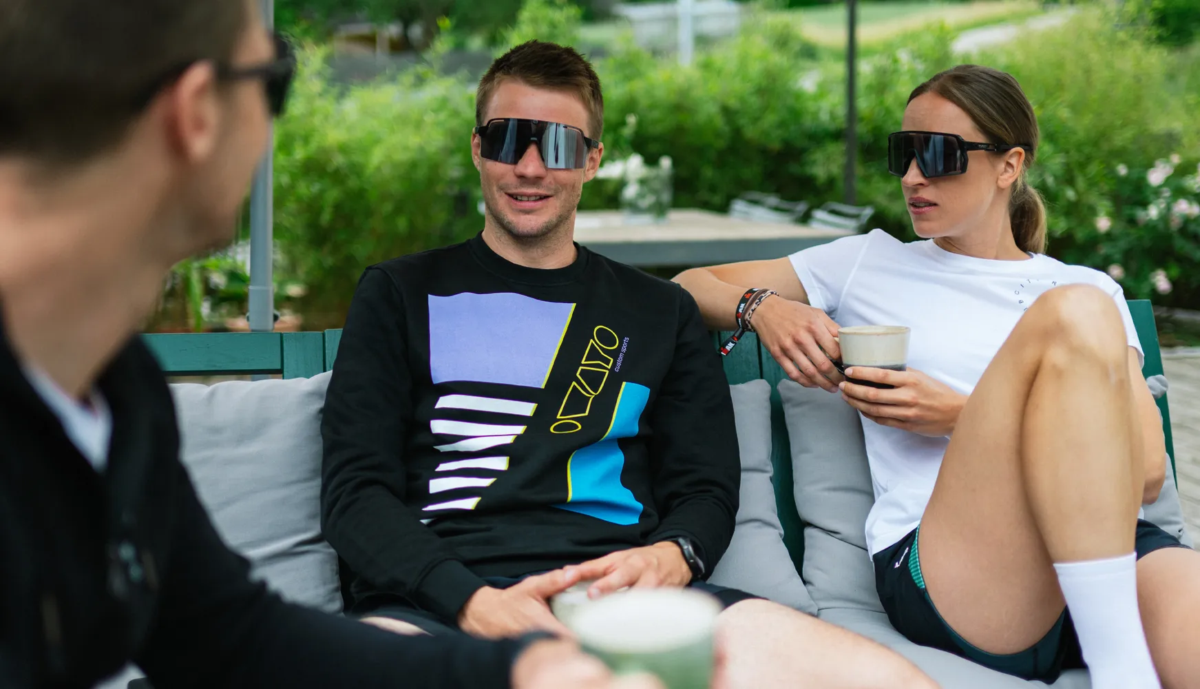 Drei Personen sitzen entspannt auf einer Couch, eine trägt einen schwarzen Pullover mit buntem Design, die andere ein weißes T-Shirt, während sie Getränke in der Hand halten.