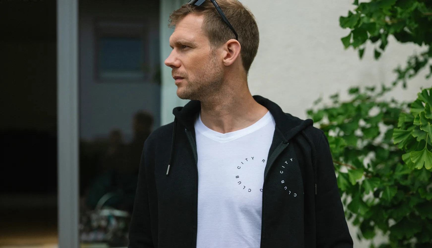 Ein Mann trägt ein schwarzes Kapuzenshirt über einem weißen T-Shirt mit einem kreisförmigen Aufdruck, während er in einem Außenbereich steht.