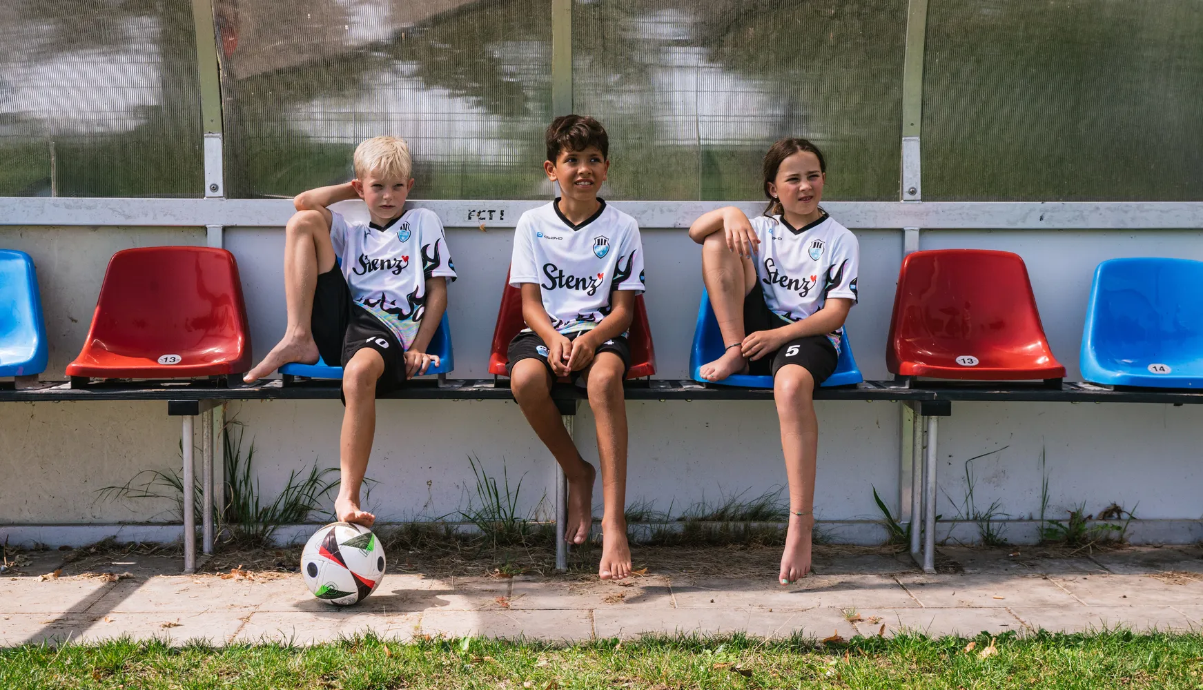 Drei Kinder in maßgeschneiderten Fußballtrikots sitzen auf einer Bank, während ein Fußball auf dem Boden liegt.