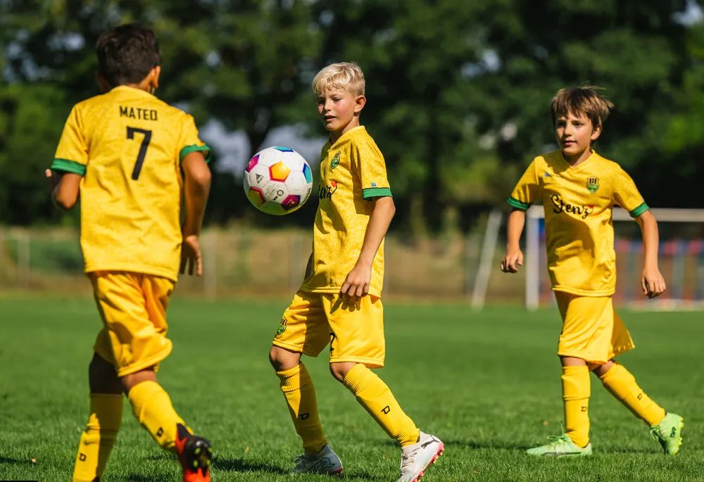 Drei Kinder in leuchtend gelben Fußballtrikots spielen auf einem Rasenfeld.