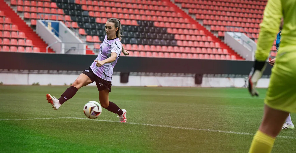 Eine Spielerin in einem lila-weißen Fußballtrikot tritt den Ball auf einem Stadionrasen, während ein Torwart in gelber Ausrüstung im Hintergrund steht.