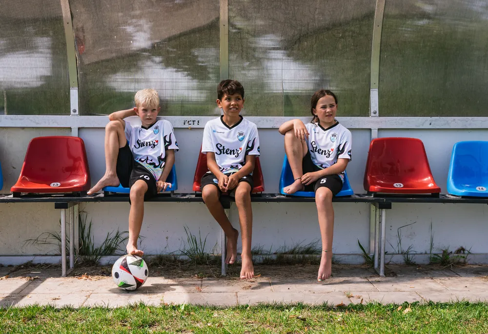 Drei Kinder in sportlichen Fußballtrikots sitzen auf einer Bank neben einem Fußball.