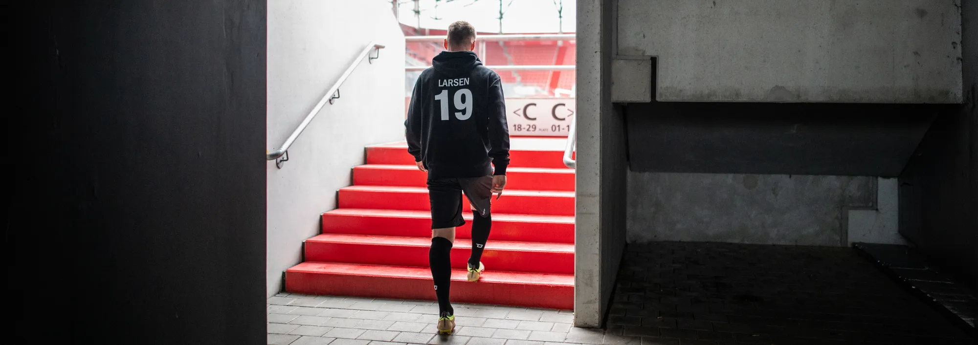 Ein Spieler in einem dunklen Kapuzenpullover und kurzen Hosen geht die roten Treppen aus einem Stadion hinauf.