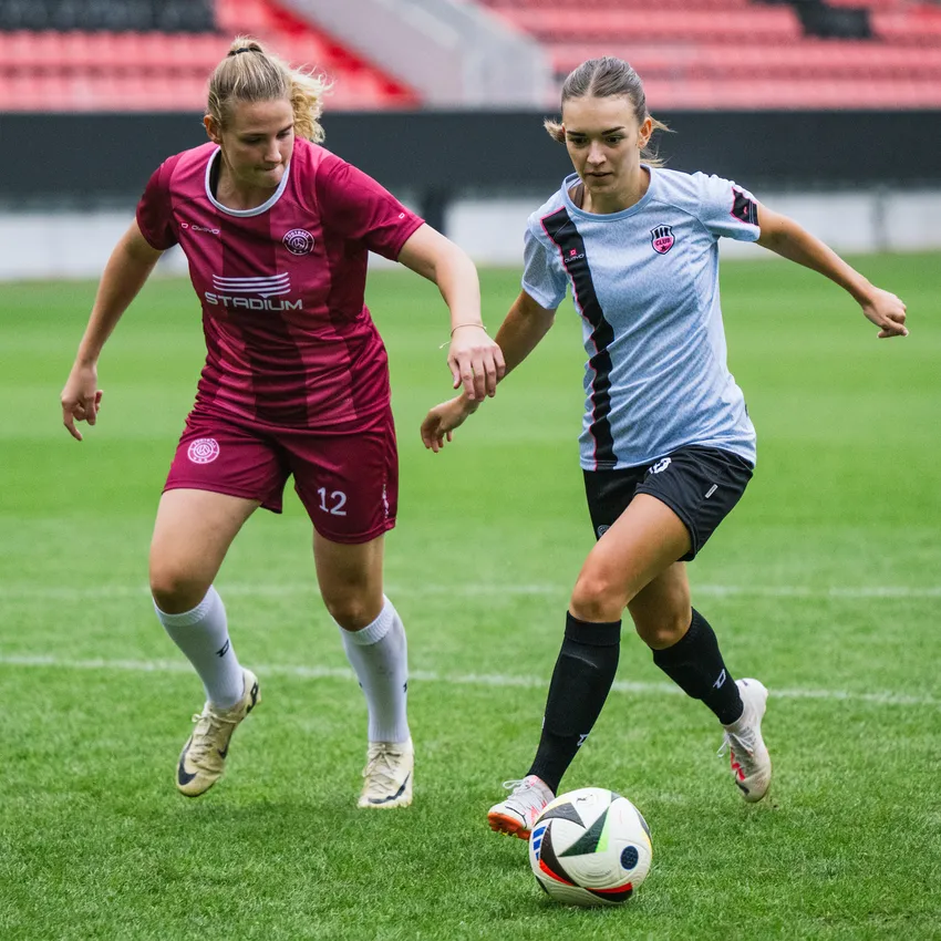 Zwei Frauen im Fußballtrikot spielen auf einem Rasenfeld und versuchen, den Ball zu kontrollieren.