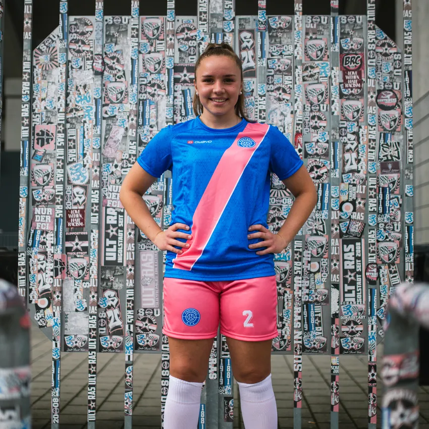 Ein Mädchen steht lächelnd vor einem Wand mit Aufklebern, während es ein blaues Fußballtrikot mit pinkem Streifen und pinken Shorts trägt.