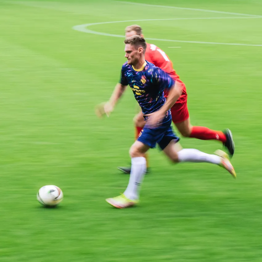 Ein Fußballspieler in einem bunten Trikot sprintet über das Spielfeld, während ein weiterer Spieler ihn verfolgt.
