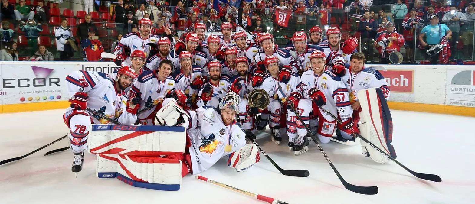 Ein Eishockeyteam in weißen Trikots mit roten und blauen Akzenten posiert auf dem Eis, umgeben von Fans, während die Spieler Medaillen tragen und einen Pokal halten.