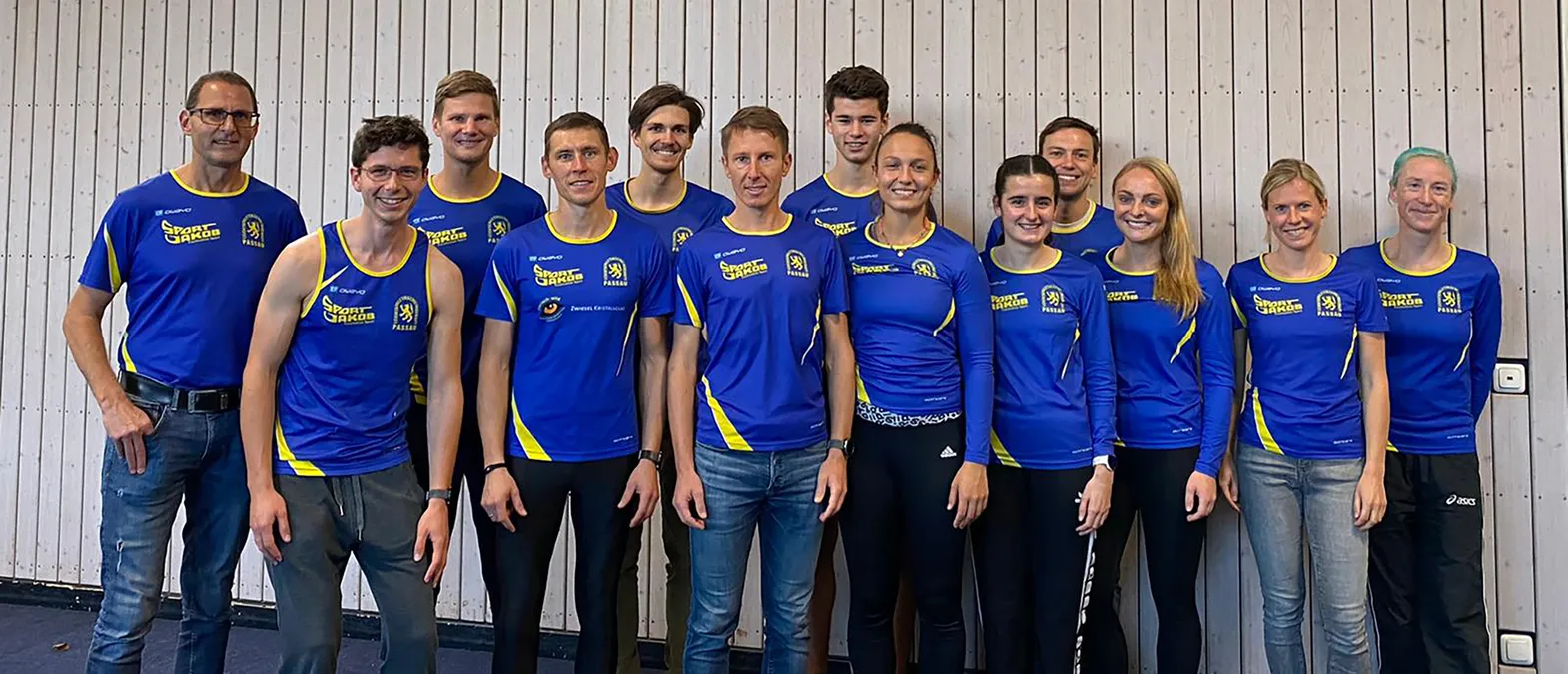 Eine Gruppe von Männern und Frauen in blauen, gelb akzentuierten Sporttrikots mit dem Logo 'Sport Jakob Passau' posiert vor einer hellen Holzwand.
