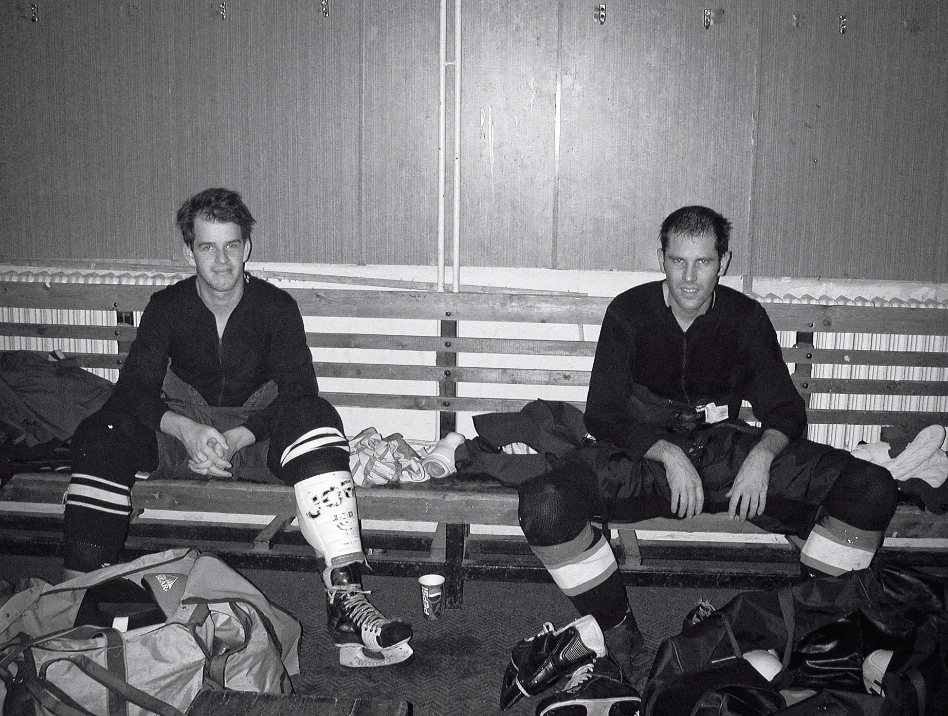Zwei Eishockeyspieler in voller Ausrüstung sitzen in einer Umkleidekabine auf einer Holzbank, umgeben von Sporttaschen und Schlägern.