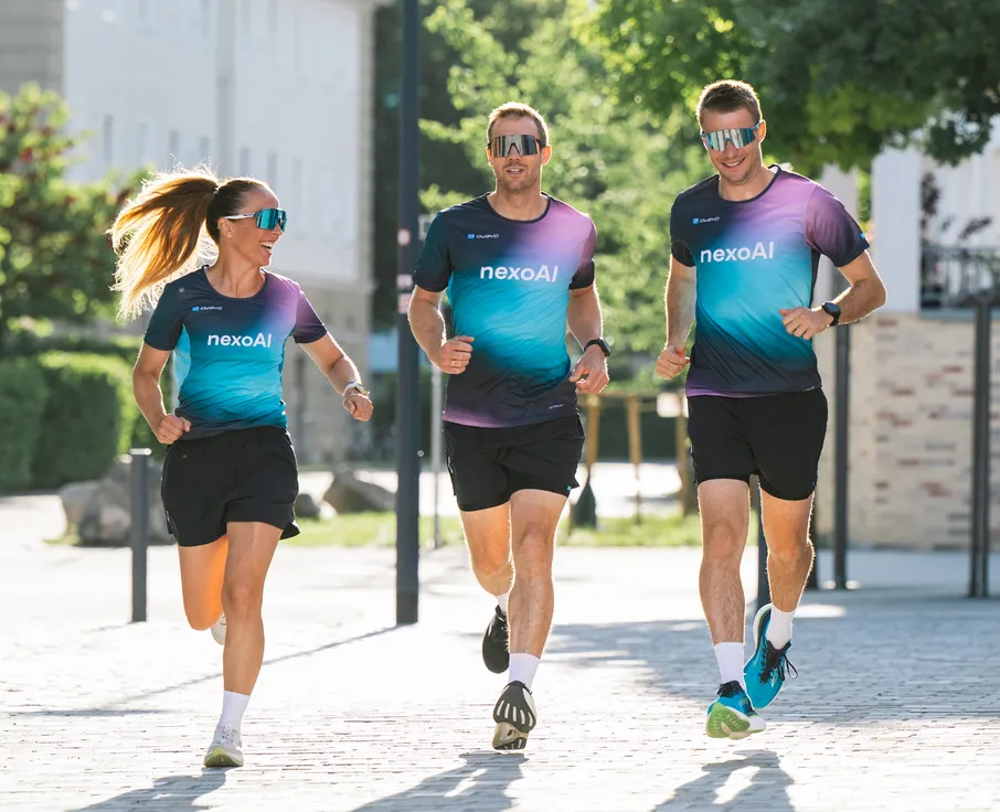 Drei Sportler, zwei Männer und eine Frau, joggen in einheitlichen, farbigen Laufshirts und Shorts auf einem gepflasterten Weg.