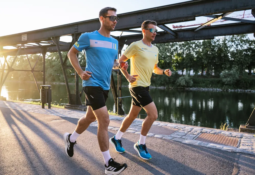 Zwei Männer in Sportbekleidung joggen entlang eines ruhigen Gewässers, während die Sonne aufgeht.