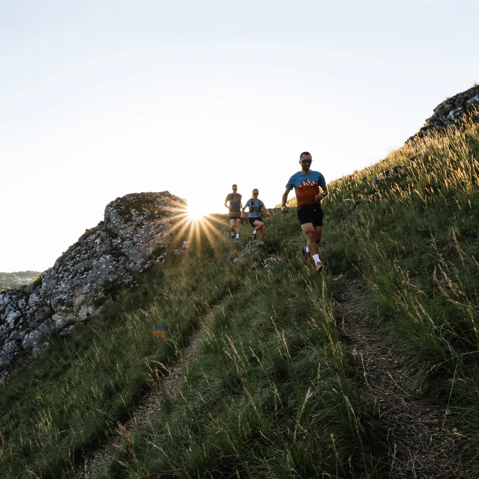 Drei Läufer in sportlicher Kleidung joggen einen hügeligen Pfad entlang, während die Sonne hinter ihnen aufgeht.