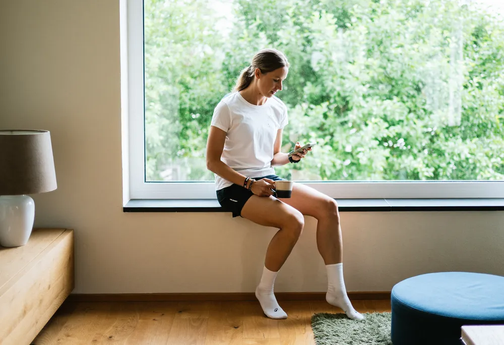 Eine Frau sitzt auf einer Fensterbank mit einem Handy in der Hand und einer Tasse in der anderen, während sie in einem weißen T-Shirt und kurzen schwarzen Shorts gekleidet ist.