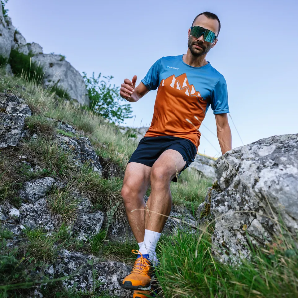 Ein Mann läuft auf einem felsigen Weg und trägt ein kurzärmeliges, blau-orangefarbenes Sporttrikot mit Bergmotiv und schwarze Shorts.