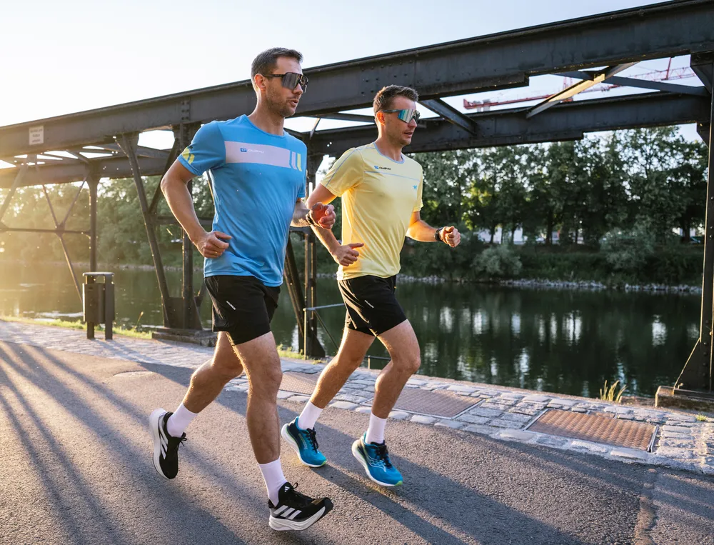 Zwei Männer joggen am Ufer eines Gewässers, einer trägt ein blaues T-Shirt und der andere ein gelbes, beide in kurzen Hosen.