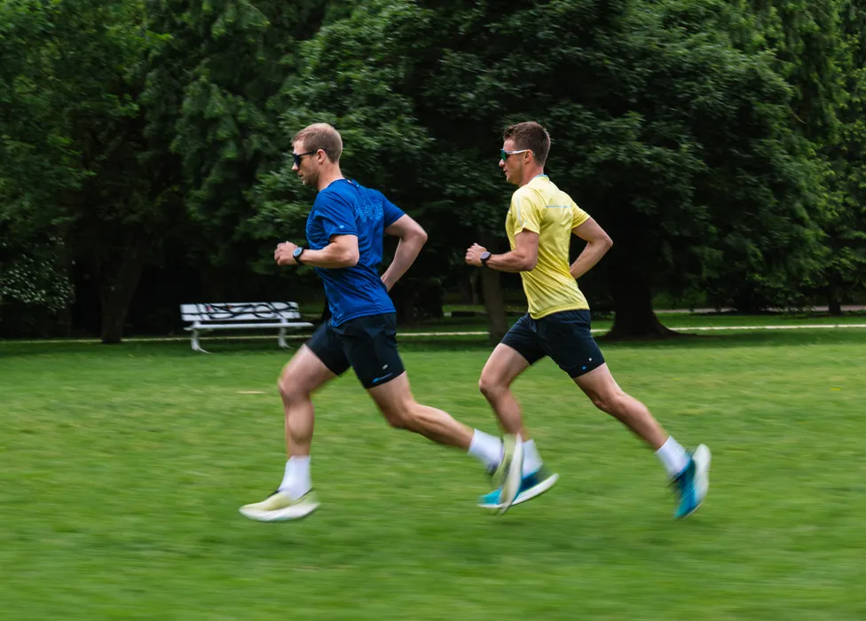 Zwei Männer in Sportkleidung rennen auf einer grünen Wiese, einer trägt ein blaues Shirt und der andere ein gelbes Shirt.