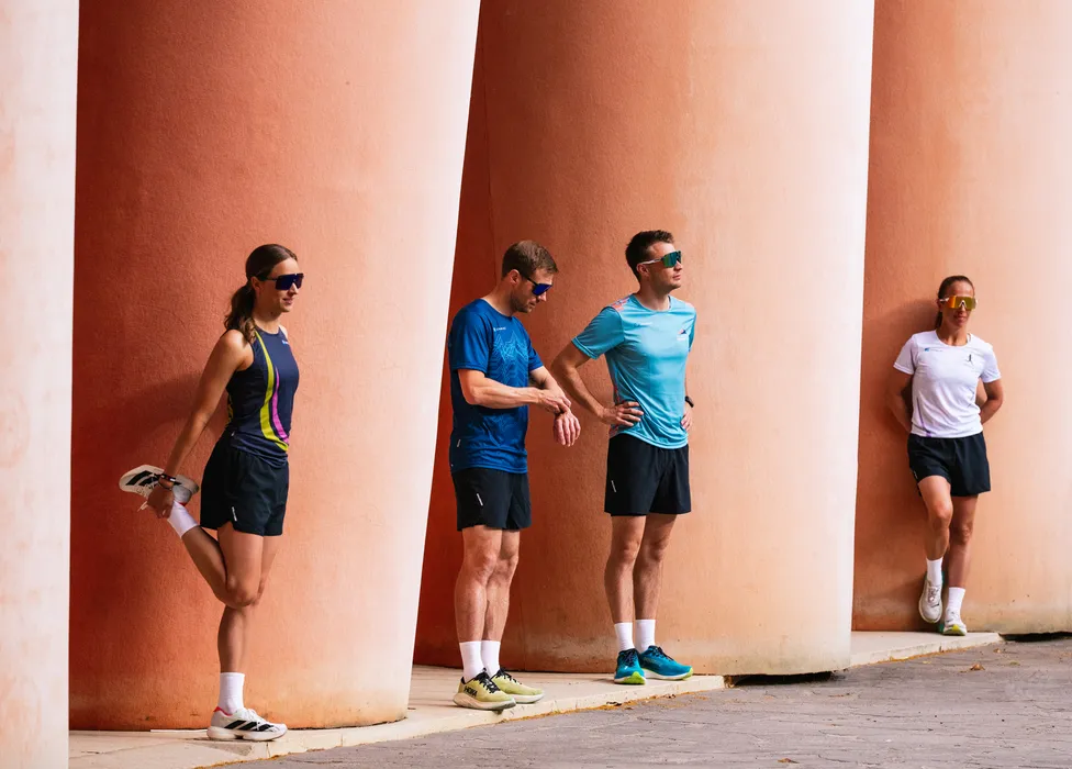 Eine Gruppe von vier Sportlern in Laufbekleidung dehnt sich an einer wand mit großen Säulen.