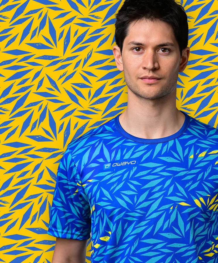 Ein junger Mann trägt ein sportliches, blaues T-Shirt mit einem bunten Blattmuster vor einem gelben Hintergrund.