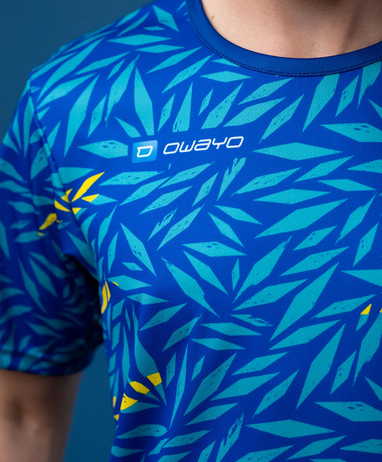 Ein sportliches, kurzärmeliges Shirt mit einem blauen, blätterartigen Muster in verschiedenen Grüntönen und gelben Akzenten.