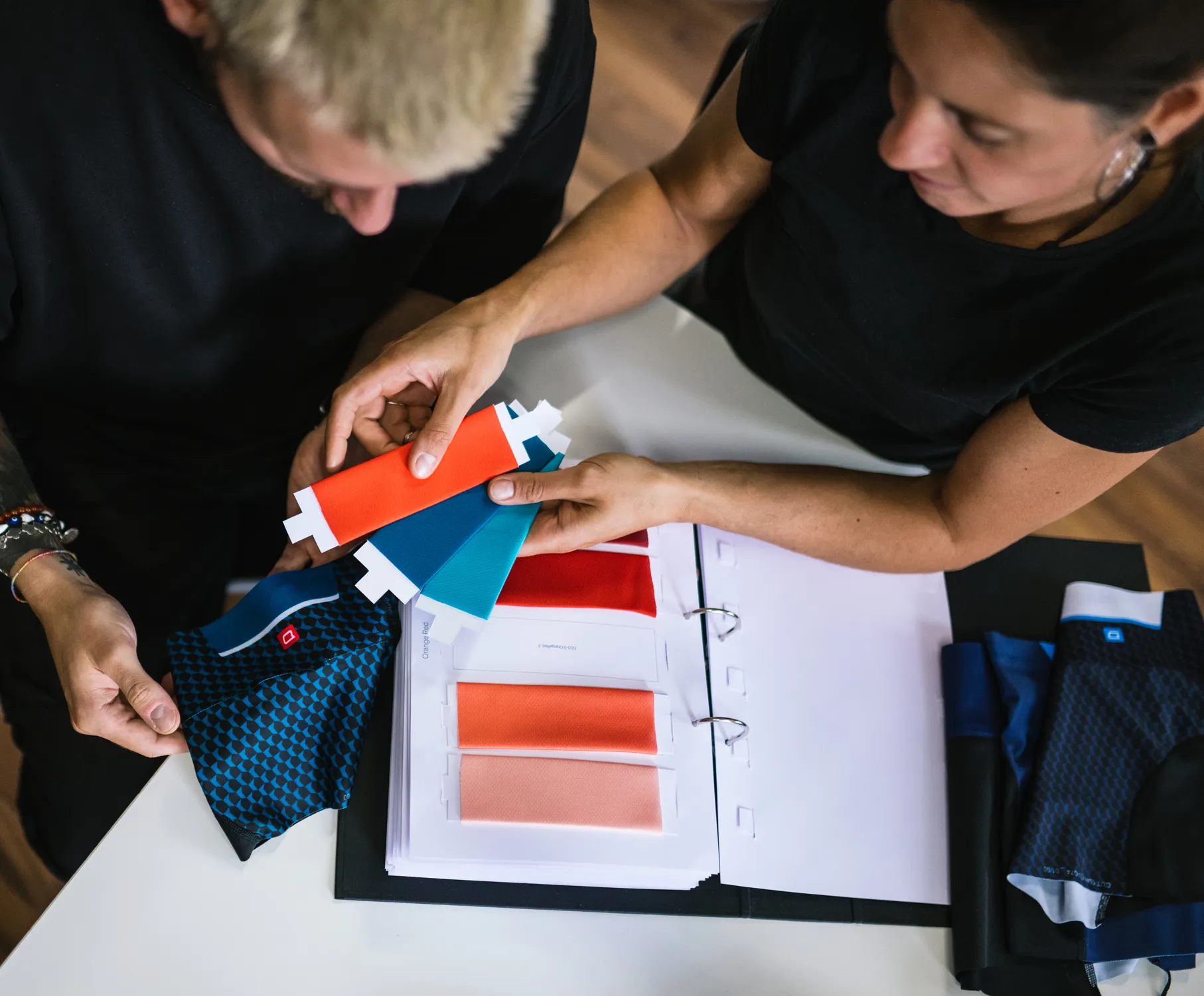 Zwei Personen wählen Farben für maßgeschneiderte Sportbekleidung aus, indem sie Stoffproben und ein Farbregister betrachten.