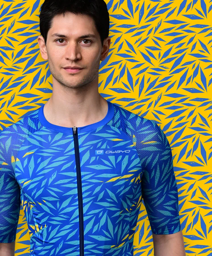 Ein Mann trägt ein blaues, kurzärmeliges Sporttrikot mit einem blauen und gelben Blattmuster, das an eine tropische Pflanze erinnert, vor einem gelben Hintergrund mit blauen Blattmustern.
