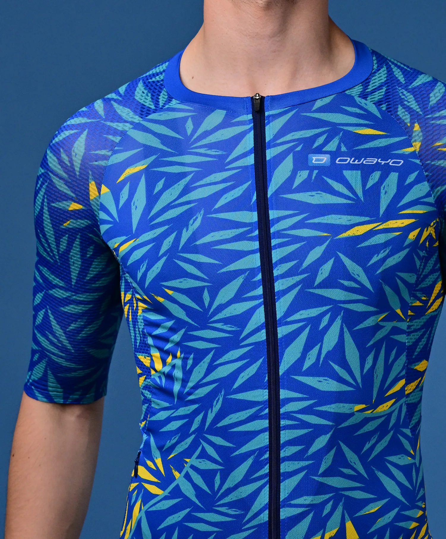 Ein Mann trägt ein blaues, kurzärmeliges Sporttrikot mit einem tropischen Blattmuster in Türkis und Gelb sowie einem Reißverschluss vorne.