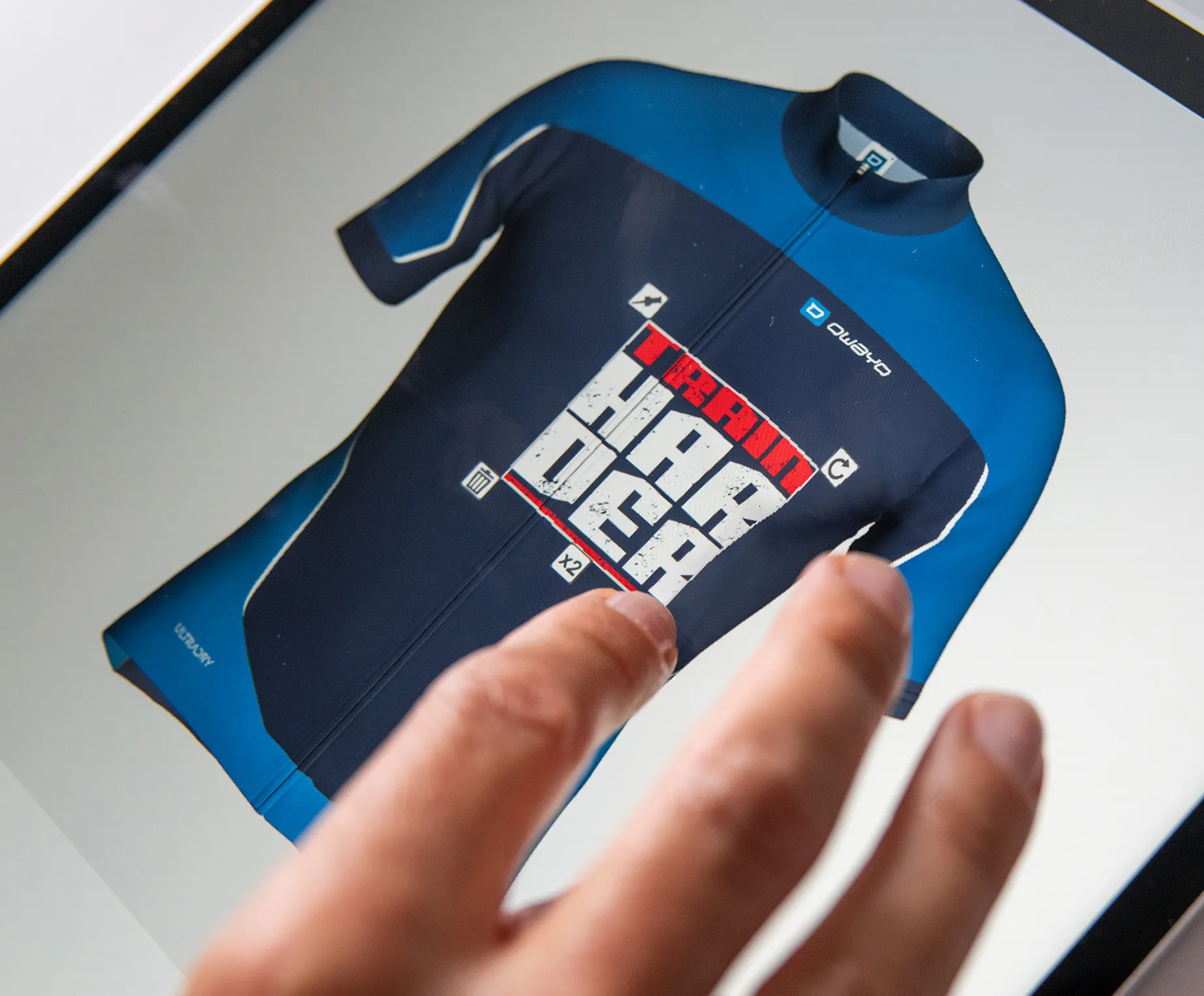 Eine Hand berührt einen Bildschirm, auf dem ein maßgeschneidertes, dunkelblaues und blaues Sporttrikot mit dem Text 'TRAIN HARDER' und dem Logo von owayo angezeigt wird.
