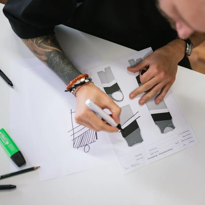 Eine Person skizziert Designs für Sportbekleidung auf Papier, umgeben von Stiften und Markern.