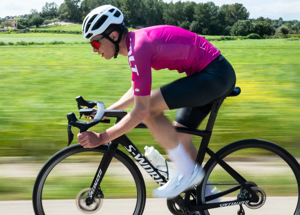 Ein Radfahrer in einem pinken Trikot und schwarzen Shorts fährt auf einer Straße durch eine grüne Landschaft, wobei die Bewegungsunschärfe die Geschwindigkeit betont.