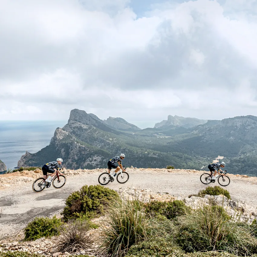 Drei Radfahrer in Sportkleidung fahren auf einer kurvenreichen Straße mit Berglandschaft und Meer im Hintergrund.