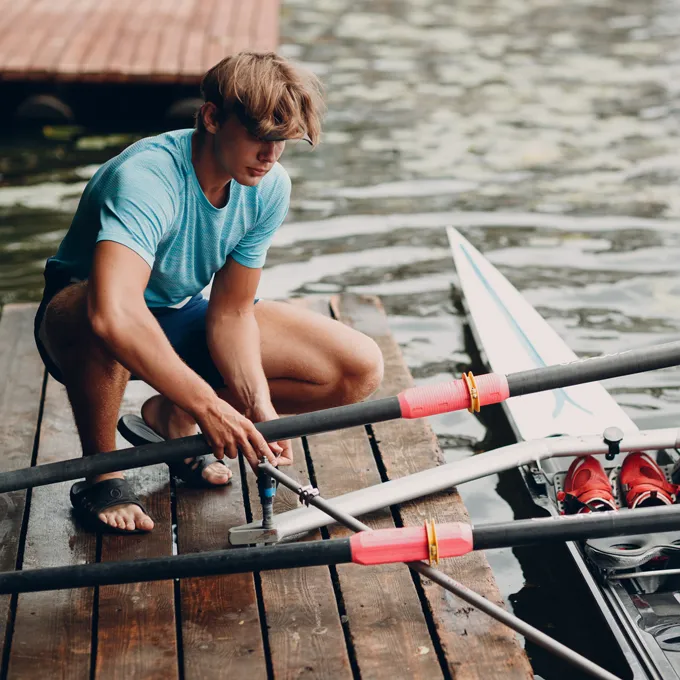 Ein junger Mann in einem blauen Sportshirt repariert Ruder auf einem Steg am Wasser.