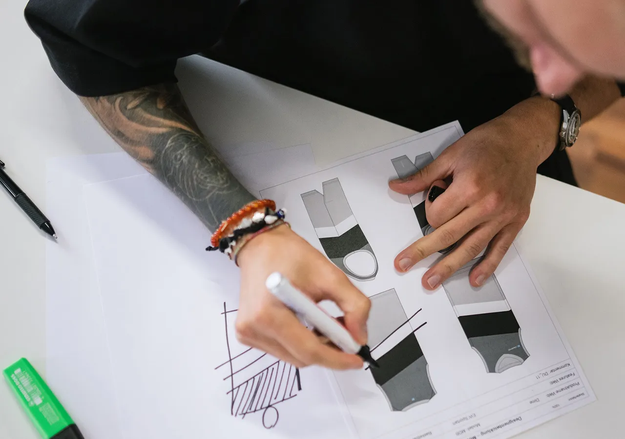Eine Person mit einem Tattoo am Arm zeichnet mit einem weißen Marker auf einem Blatt Papier, auf dem mehrere Entwürfe von sportlichen Kleidungsstücken zu sehen sind.