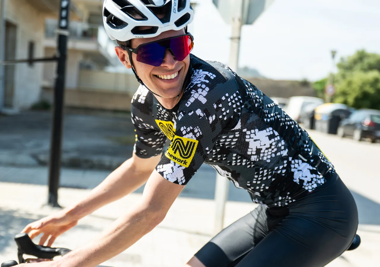 Ein Radfahrer in einer schwarzen und weißen, gemusterten Radtrikot mit einem gelben Sponsorenlogo, der auf einer Straße fährt.
