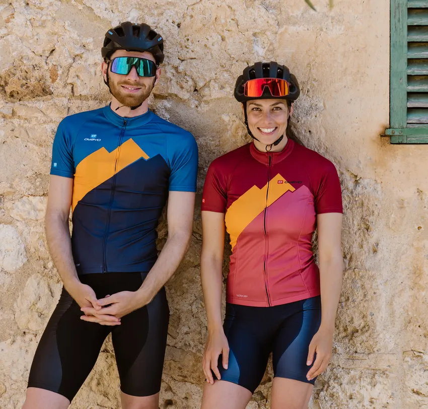 Ein Mann und eine Frau in maßgeschneiderten Radtrikots von owayo, die vor einer Steinmauer stehen.